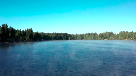 Lake-fog-rises-from-blue-water-surrounded-by-pine-tree-forest,-drone-dolly-push-in