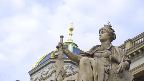 woman-statue-sitting-down-on-a-throne-holding-a-sword-in-front-of-her-wearing-a-crown-with-a-cross-at-the-background-a-building-with-golden-tower-pointed-top-and-details-made-by-professional