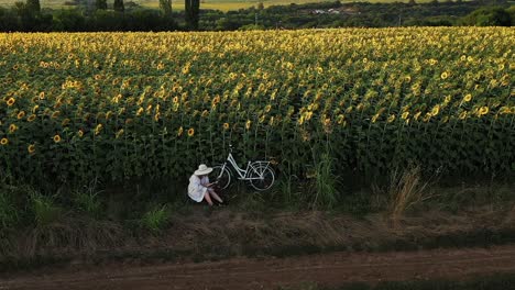 Girl-in-white-dress-reads-book-in-sunflower-field-golden-hour-drone-push-shot
