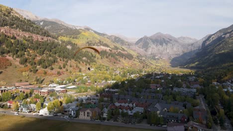A-rotating-drone-shot,-of-a-paraglider-flying-over-a-neighborhood-in-the-town-of-Telluride,-Colorado,-on-a-sunny-day-in-the-Fall-season