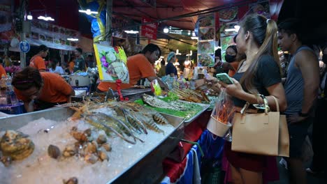 Auf-Der-Rechten-Seite-Sind-Kunden-Zu-Sehen,-Die-Auf-Dem-Chatuchak-Wochenendnachtmarkt-In-Bangkok,-Thailand,-Auf-Ihre-Bestellungen-Von-Meeresfrüchten-Und-Schweine--Und-Hähnchengrill-Von-Lokalen-Anbietern-Warten