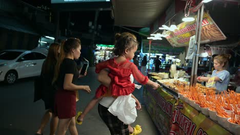 Drei-Frauen-Und-Ein-Baby-Möchten-Obst-Und-Snacks-Von-Freunden-Kaufen,-Die-In-Einem-Imbissstand-Auf-Dem-Chatuchak-Wochenendnachtmarkt-In-Bangkok,-Thailand,-Ausgestellt-Sind