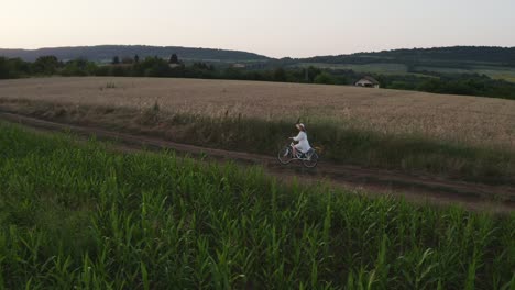 Lone-girl-in-white-dress-rides-bike-through-rural-farmland-at-golden-hour