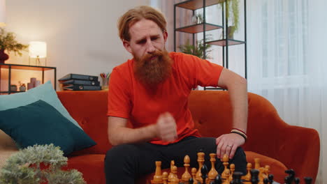 Focused-bearded-redhead-young-man-playing-chess-leisure-board-game-alone,-domestic-activity-at-home