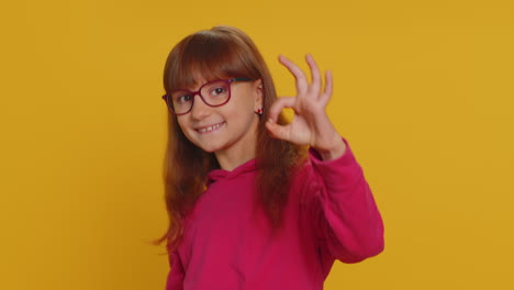 Young-toddler-school-girl-in-glasses-looking-approvingly-at-camera-showing-okey-gesture,-like-sign