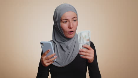Feliz-Mujer-Musulmana-Usa-Un-Teléfono-Inteligente-Regocijándose-Por-Ganar,-Recibiendo-Dinero-En-Efectivo-En-Dólares,-Suerte-De-Lotería-De-éxito