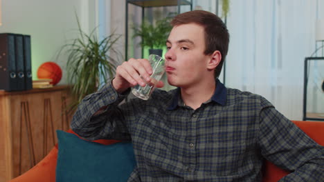 Thirsty-teen-young-man-sitting-at-home-holding-glass-of-natural-aqua-make-sips-drinking-still-water