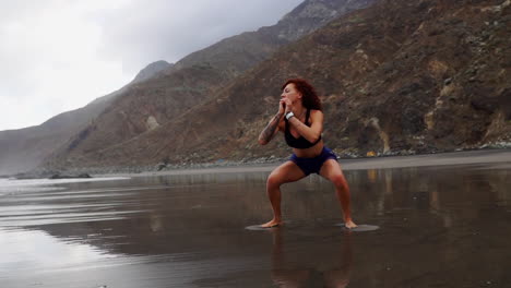 Slow-motion-captures-a-young,-slender-woman-engaging-in-squats-and-jumps-for-her-training-by-the-ocean's-shore