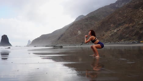 Performing-squats-and-jumps-by-the-ocean's-shore,-a-young,-slim-woman's-training-is-depicted-in-slow-motion
