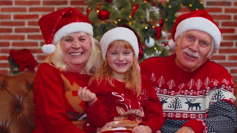 Senior-grandparents-with-granddaughter-in-Santa-hats-laughing-out-loud-at-home-near-Christmas-tree
