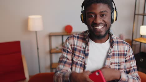 Pretty-African-American-man-in-headphones-listening-music-dancing,-singing-in-living-room-at-home