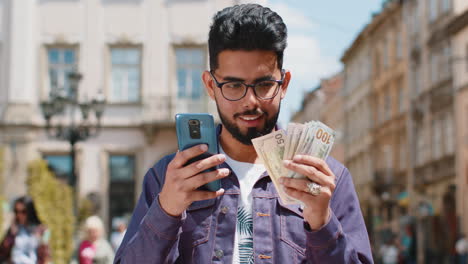 Fröhlicher-Junger-Indischer-Mann,-Der-Geld-Zählt,-Dollar-Bargeld-Verwendet-Smartphone-Rechner-App-In-Der-Stadtstraße