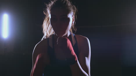 A-beautiful-female-boxer-strikes-directly-into-the-camera-looking-into-the-camera-and-moving-forward-on-a-dark-background-with-a-backlight.-Steadicam-shot