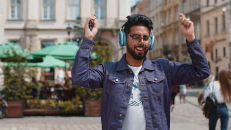 Happy-young-indian-man-in-wireless-headphones,-listening-favorite-energetic-music-in-city-street