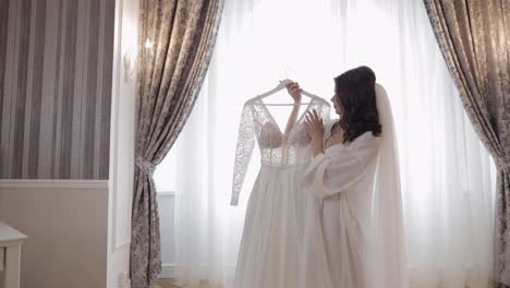 Braut-In-Dessous-Steht-Mit-Ihrem-Hochzeitskleid-Zu-Hause-Am-Fenster,-Weißem-Boudoir-Kleid-Und-Schleier