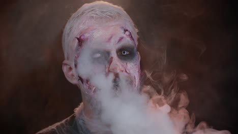 Sinister-man-horrible-scary-Halloween-zombie-looking-ominous-at-camera-blows-smoke-from-nose,-mouth