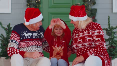Elderly-grandmother-grandfather-with-granddaughter-sitting-at-Christmas-house-porch-waving-hello-hi