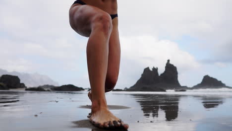 A-female-athlete-does-slow-motion-exercises,-stretching-her-legs-and-doing-lunges-at-the-beach,-while-a-fitness-woman-practices-calming-exercises-facing-the-sea