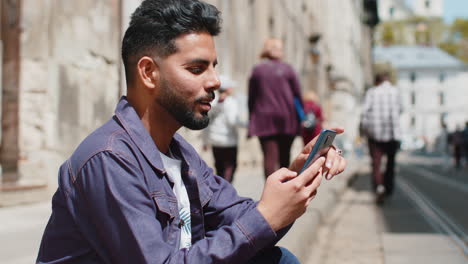 Indischer-Mann-Benutzt-Smartphone,-Schreibt-Textnachrichten,-Surft-Im-Internet-Und-Beendet-Seine-Arbeit-Mit-Blick-In-Die-Kamera