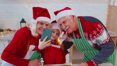 Grandparents-family-with-granddaughter-kid-taking-selfie-photo-on-mobile-phone-at-Christmas-kitchen