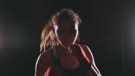 A-beautiful-female-boxer-strikes-directly-into-the-camera-looking-into-the-camera-and-moving-forward-on-a-dark-background-with-a-backlight.-Steadicam-shot