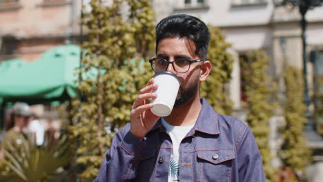 Young-indian-man-enjoying-drinking-morning-coffee-hot-drink,-relaxing,-taking-a-break-in-city-street