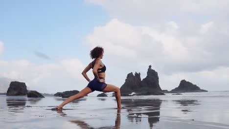 In-slow-motion,-a-young-woman-stands-by-the-ocean's-edge,-engaging-in-stretching-and-yoga-while-gazing-into-the-distance,-embodying-serenity
