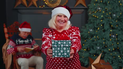 Ältere-Großmutter-Im-Neujahrspullover-Und-Hut-Präsentiert-Weihnachtsgeschenkbox,-Feiertage-Zu-Hause