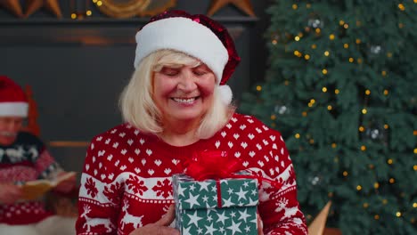 Elderly-grandmother-in-festive-sweater-presenting-Christmas-gift-box,-smiling,-looking-at-camera