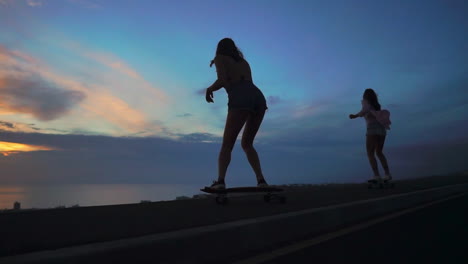 In-slow-motion,-two-friends-skate-along-a-road-at-sunset,-with-mountains-and-a-stunning-sky-as-the-backdrop.-They're-wearing-shorts