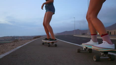 Two-friends,-attired-in-shorts,-skateboard-along-a-road-during-sunset,-against-the-backdrop-of-mountains-and-a-beautiful-sky,-all-captured-in-slow-motion