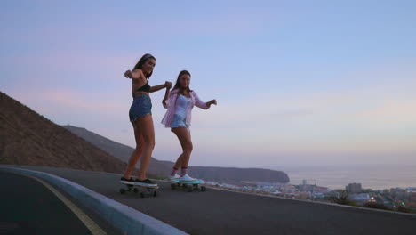 Zwei-Freunde-In-Kurzen-Hosen-Fahren-Bei-Sonnenuntergang-Langsam-Auf-Einer-Straße-Skateboard,-Mit-Blick-Auf-Berge-Und-Einen-Faszinierenden-Himmel