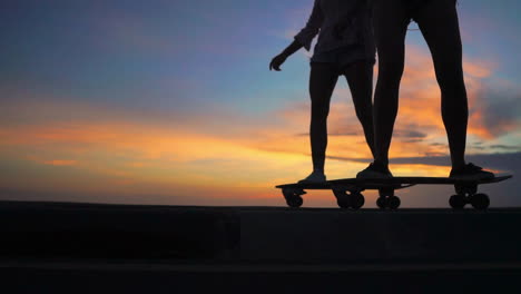 Vor-Der-Kulisse-Von-Bergen-Und-Einem-Wunderschönen-Himmel-Fahren-Zwei-Freunde-Bei-Sonnenuntergang-Skateboards-Auf-Einer-Straße,-Aufgenommen-In-Zeitlupe.-Sie-Tragen-Kurze-Hosen