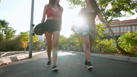 Amid-palm-trees,-girlfriends-walk-to-the-park,-carrying-skateboards,-engaging-in-conversation,-laughter,-and-joyful-smiles-in-the-sunset's-warm-hues,-all-in-slow-motion