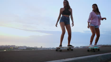 In-slow-motion,-two-friends-wearing-shorts-ride-skateboards-along-a-road-at-sunset,-with-mountains-and-a-captivating-sky-as-the-backdrop