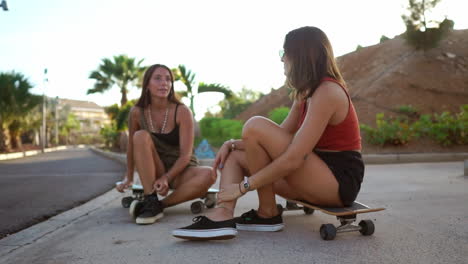 With-the-sun-setting,-two-girls-in-a-skate-park-relax-on-their-boards,-exchanging-conversation,-accompanied-by-smiles-and-hearty-laughter.-Enjoying-a-longboard-talk-among-friends