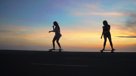 As-the-sun-sets,-two-friends-skateboard-on-a-road-with-mountains-and-a-beautiful-sky-in-the-background,-all-captured-in-slow-motion.-They-sport-shorts