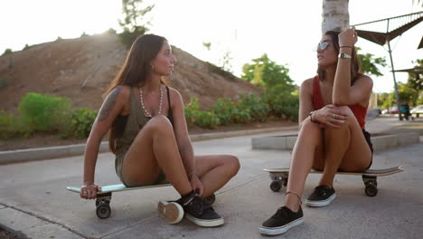 In-the-soft-light-of-the-sunset,-two-girls-on-skateboards-at-the-skate-park-engage-in-a-lively-conversation,-smiling,-and-laughter-echoing-through-the-air.-Longboarding-and-friendly-banter