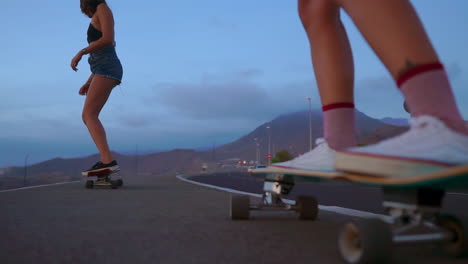In-slow-motion,-two-friends-skateboard-on-a-road-at-sunset,-with-mountains-and-a-beautiful-sky-enhancing-the-scenery.-They're-dressed-in-shorts