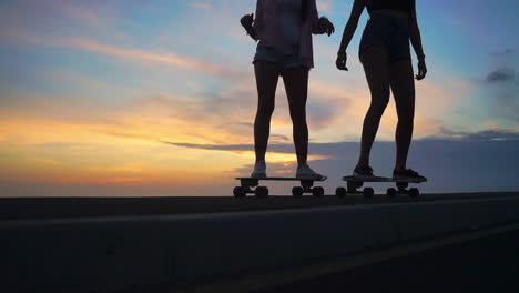 In-the-midst-of-a-captivating-sunset,-two-friends-skate-on-a-road,-framed-by-mountains-and-a-picturesque-sky-in-slow-motion.-Shorts-are-their-choice-of-clothing