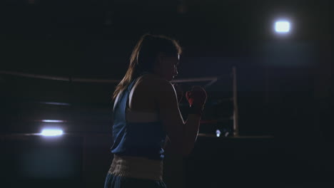 Professional-beautiful-female-boxer-otbryvatyvat-blows-conducting-a-fight-with-a-shadow-in-a-dark-hall-of-the-hall-in-slow-motion-in-blue-clothes-and-red-bandages-on-her-wrists.-flying-around-the-object.-steadicam-shot
