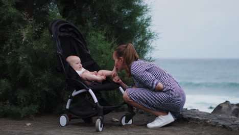 An-affectionate-and-caring-young-mother-interacts-with-her-baby-in-the-stroller,-creating-a-heartwarming-scene-for-this-sweet-young-family-against-the-ocean-backdrop