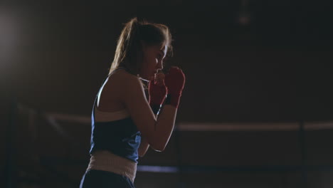 steadicam-shot-Professional-beautiful-female-boxer-unloads-strikes-while-conducting-a-shadow-fight-in-a-dark-hall-room-in-slow-motion-in-a-blue-dress-and-red-bandages-on-her-wrists.-flying-around-the-object