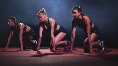 Three-sports-girls-in-black-clothes-of-the-athlete-at-night-on-the-treadmill-will-start-for-the-race-at-the-sprint-distance-from-the-sitting-position.