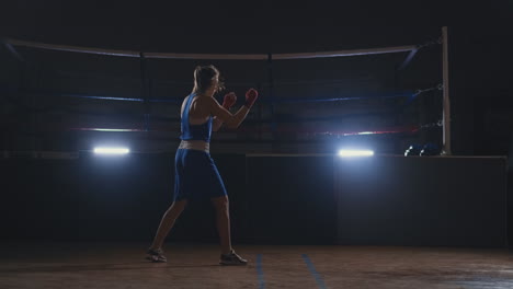 Professional-beautiful-female-boxer-shoots-off-conducting-a-shadow-fight-in-a-dark-hall-room-in-slow-motion-in-blue-clothes-and-red-bandages-on-her-wrists.-steadicam-shot