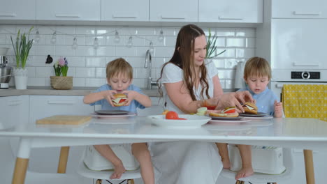 Eine-Glückliche-Familie-Ist-Eine-Junge-Schöne-Mutter-In-Einem-Weißen-Kleid-Mit-Zwei-Söhnen-In-Blauen-Hemden,-Die-Gemeinsam-Eine-Weiße-Küche-Vorbereiten,-Gemüse-Schneiden-Und-Gesundes-Berger-Für-Kinder-Zubereiten.