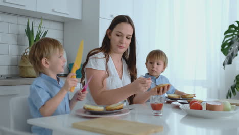 A-happy-family-is-a-young-beautiful-mother-in-a-white-dress-with-two-sons-in-blue-shirts-preparing-a-white-kitchen-together-slicing-vegetables-and-creating-healthy-berger-for-children.