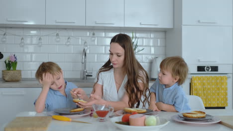 Young-mother-with-two-young-sons-in-the-kitchen-at-the-table-preparing-burger-for-lunch