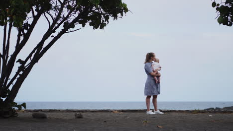 A-picturesque-portrait-captures-a-mother's-kiss-shared-with-her-daughter-by-the-sea-and-beach.-A-content-family-on-vacation,-the-mom-envelops-her-toddler-in-a-warm-embrace