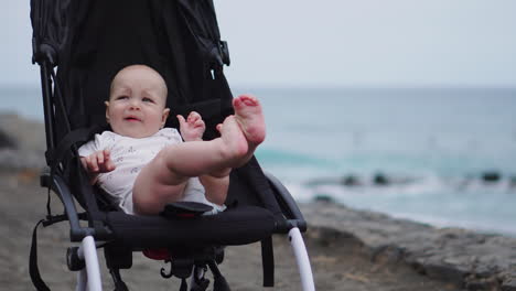 On-a-sandy-beach,-a-baby-in-a-stroller-gazes-tranquilly-into-the-boundless-expanse-of-sea-waves,-absorbed-in-the-serene-beauty-of-a-late-summer-evening-by-the-sea